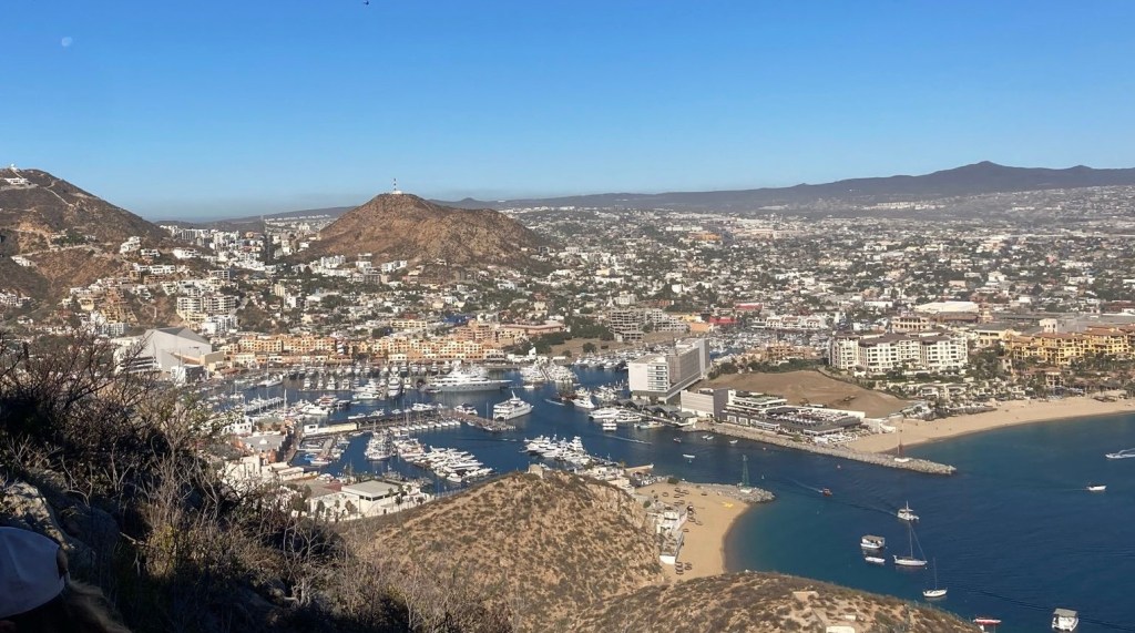 A panoramic view of a coastal city featuring a marina filled with yachts, sandy beaches, and residential buildings set against mountainous terrain under a clear blue sky.