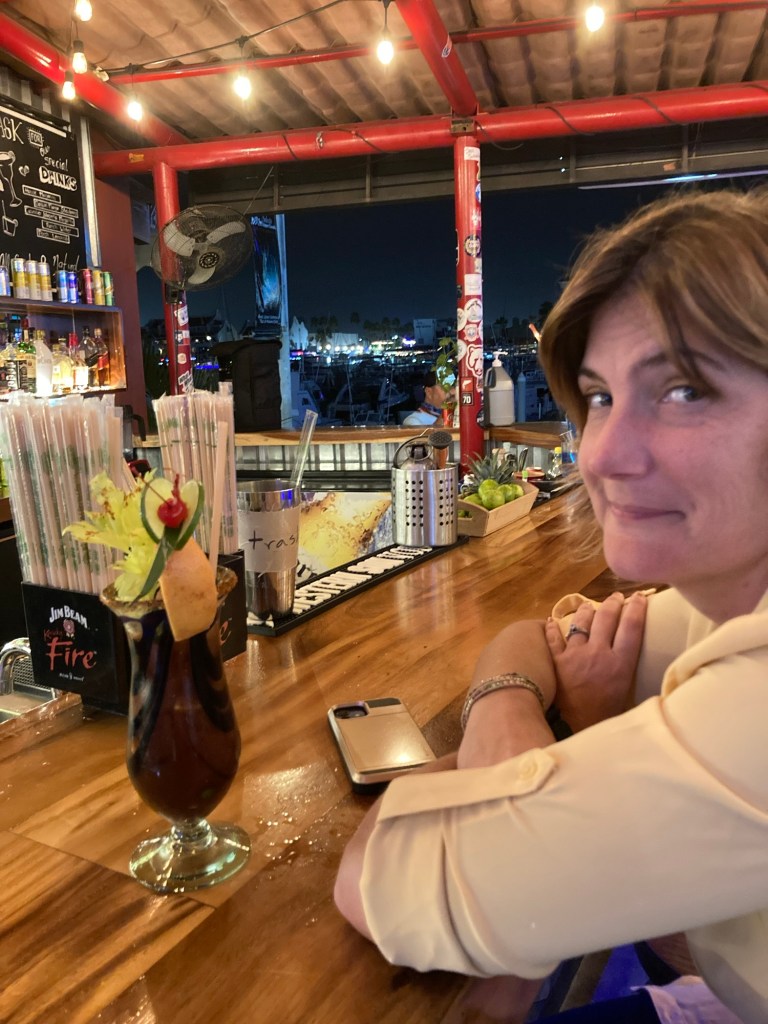 A woman sitting at a bar, looking towards the camera with a cocktail in front of her, surrounded by colorful bottles and bar decor under dim lighting.