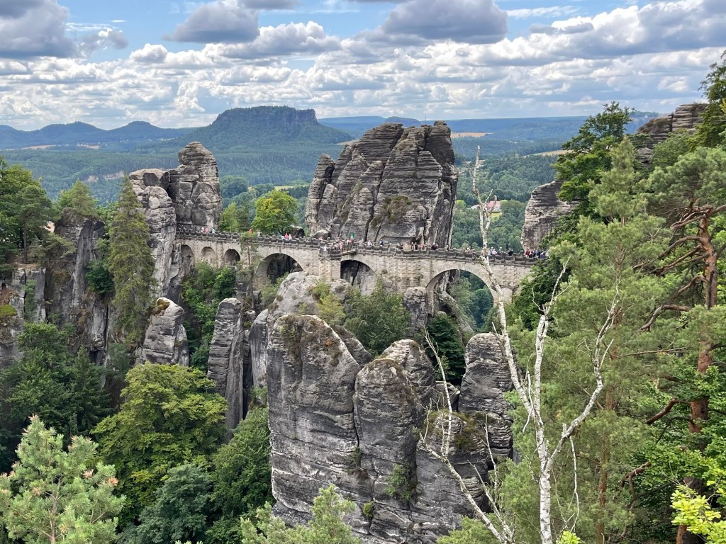 A panoramic view of a stone bridge spanning rocky formations in a lush green landscape, with distant hills under a cloudy sky.